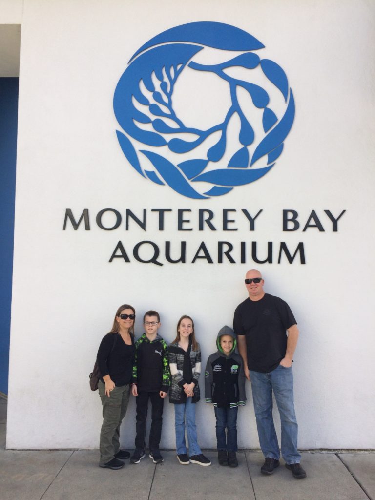 Monterey Bay Aquarium sign out front with family posing in front of it.
