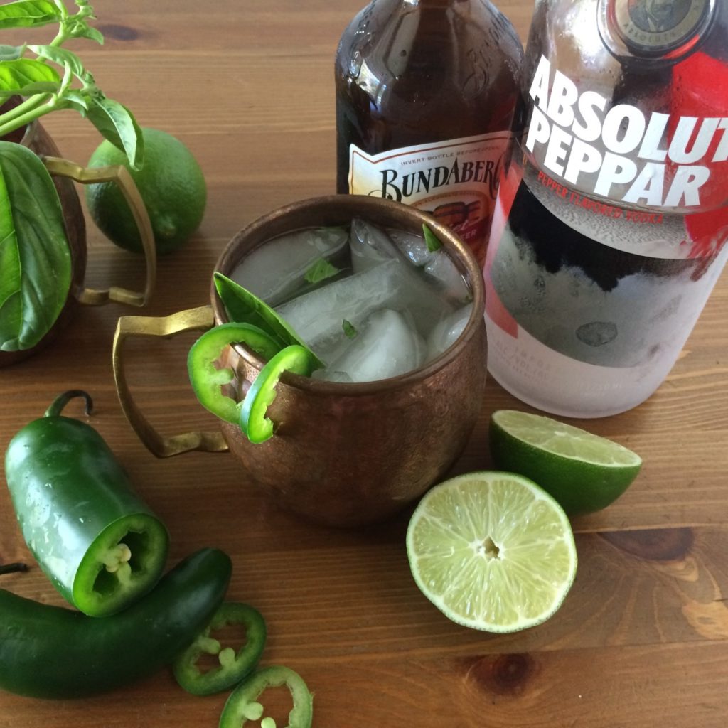 Spicy jalapeno moscow mule in copper mugs next to fresh limes absolut peppar vodka and bottles of ginger beer. 
