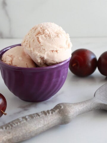 Plum Ice Cream in a bowl with fresh plums and a scoop on the table.