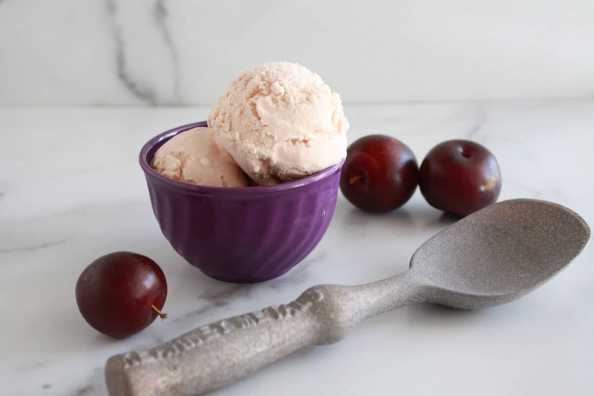 Plum Ice Cream in a bowl with fresh plums and a scoop on the table.