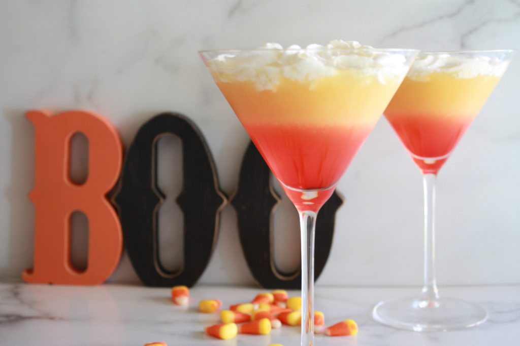 Two martini glasses filled with a colorful, layered cocktail resembling candy corn, topped with whipped cream, sit on a marble surface. In the background, the word "BOO" is spelled out in large, decorative letters, with candy corn pieces scattered nearby.