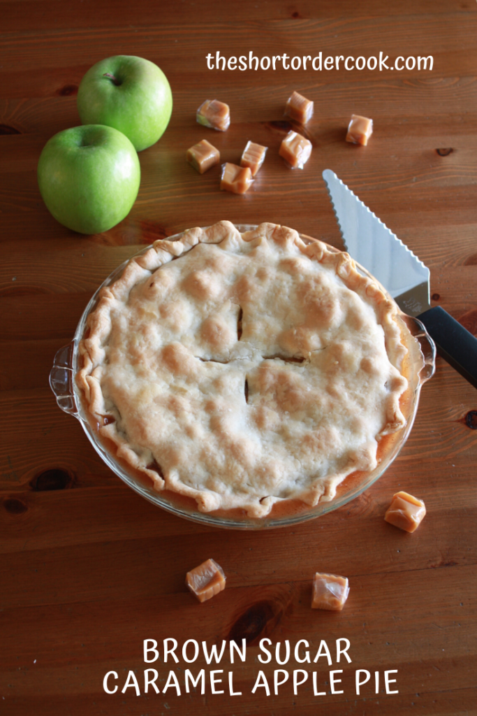 Whole apple pie on the table with a pie cutter, apples, and caramel candies on a table.