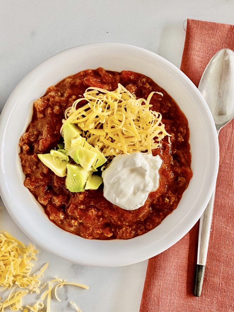 Bowl of Slow Cooker Beef &amp; Bacon Chili (no beans) topped with shredded cheddar cheese and sour cream