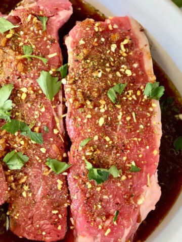 Keto Steak Marinade featured close up of 3 steaks in a white dish with marinade and fresh parsley on the table next to it