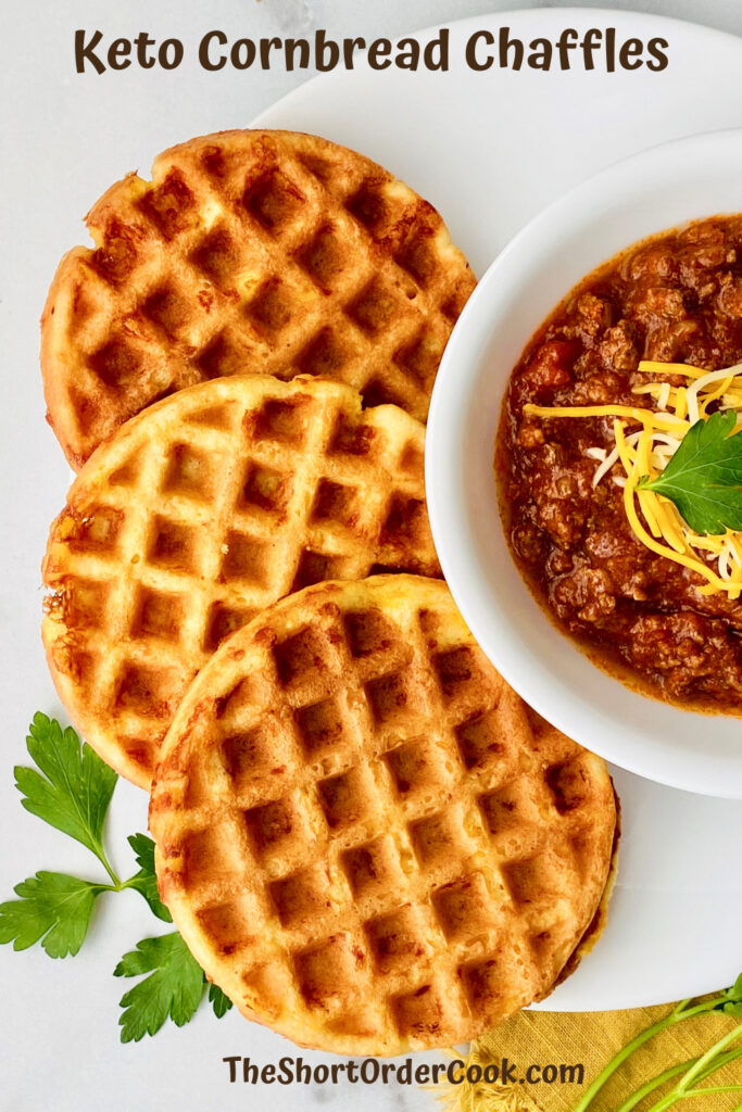 Chaffles made with almond flour to taste like cornbread fanned out on the left side and a partial bowl of chili topped with cheese on the right plus parsley sprigs on the table