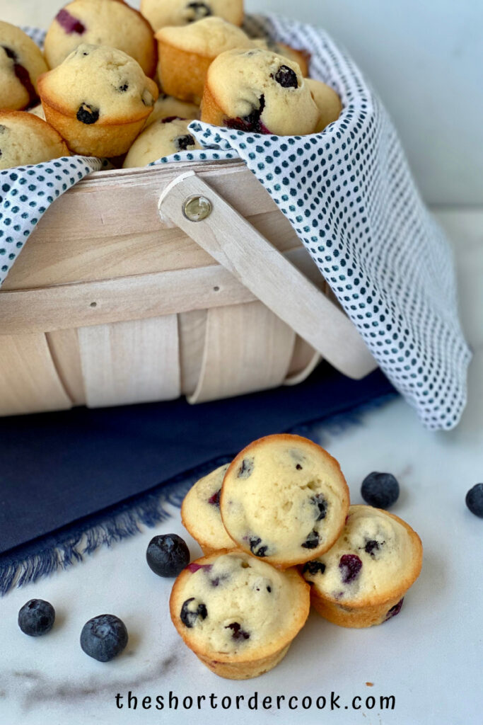 Old-Fashioned Mini Blueberry Muffins basket full and a four stacked on the side with blueberries and blue cloth napkin