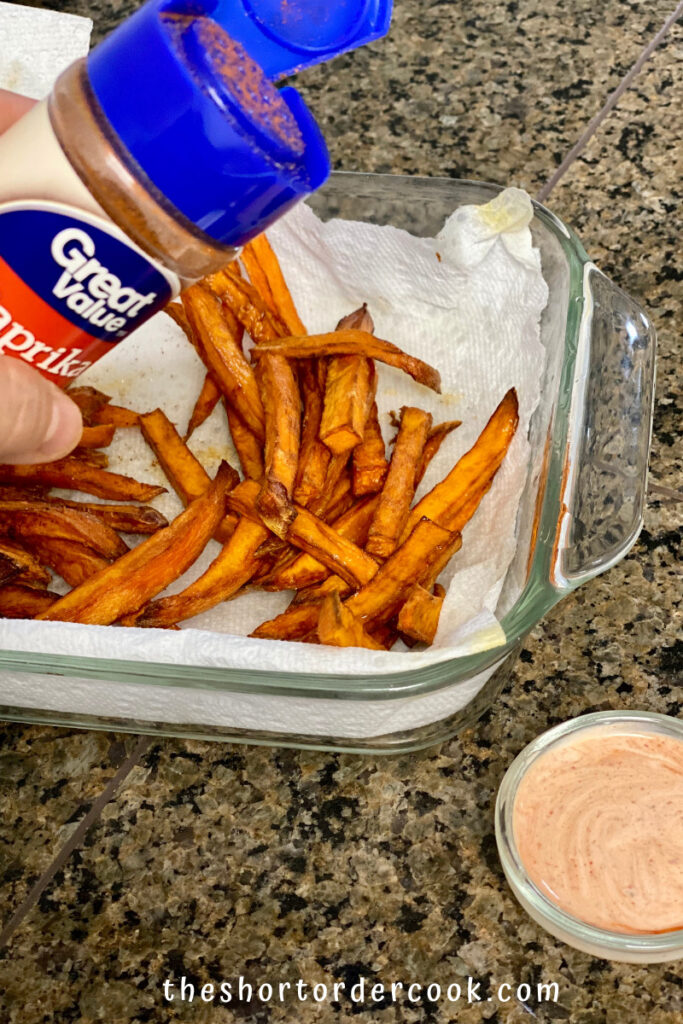 Deep Fried Sweet Potato Fries a glass dish lined with paper towel and cooked fries in it with paprika being sprinkled on them and a small glass bowl to the side with dipping sauce