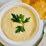Instant Pot Cream of Celery Soup featured overhead of bowl full of soup topped with parsley sprig and crackers to the side and a spoon on the right all on top a green cloth napkin recipe card image
