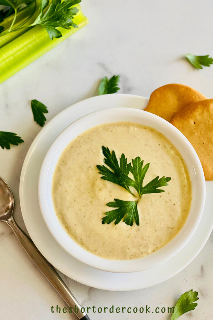 Instant Pot Cream of Celery Soup ready to eat in a bowl with crackers and a spoon on the table and celery stalks and parsley in the background
