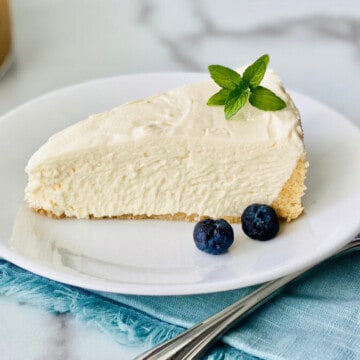 Keto No-Bake Cheesecake recipe card slice on a plate with mint and blueberries fork and napkin and whole cheesecake in the background