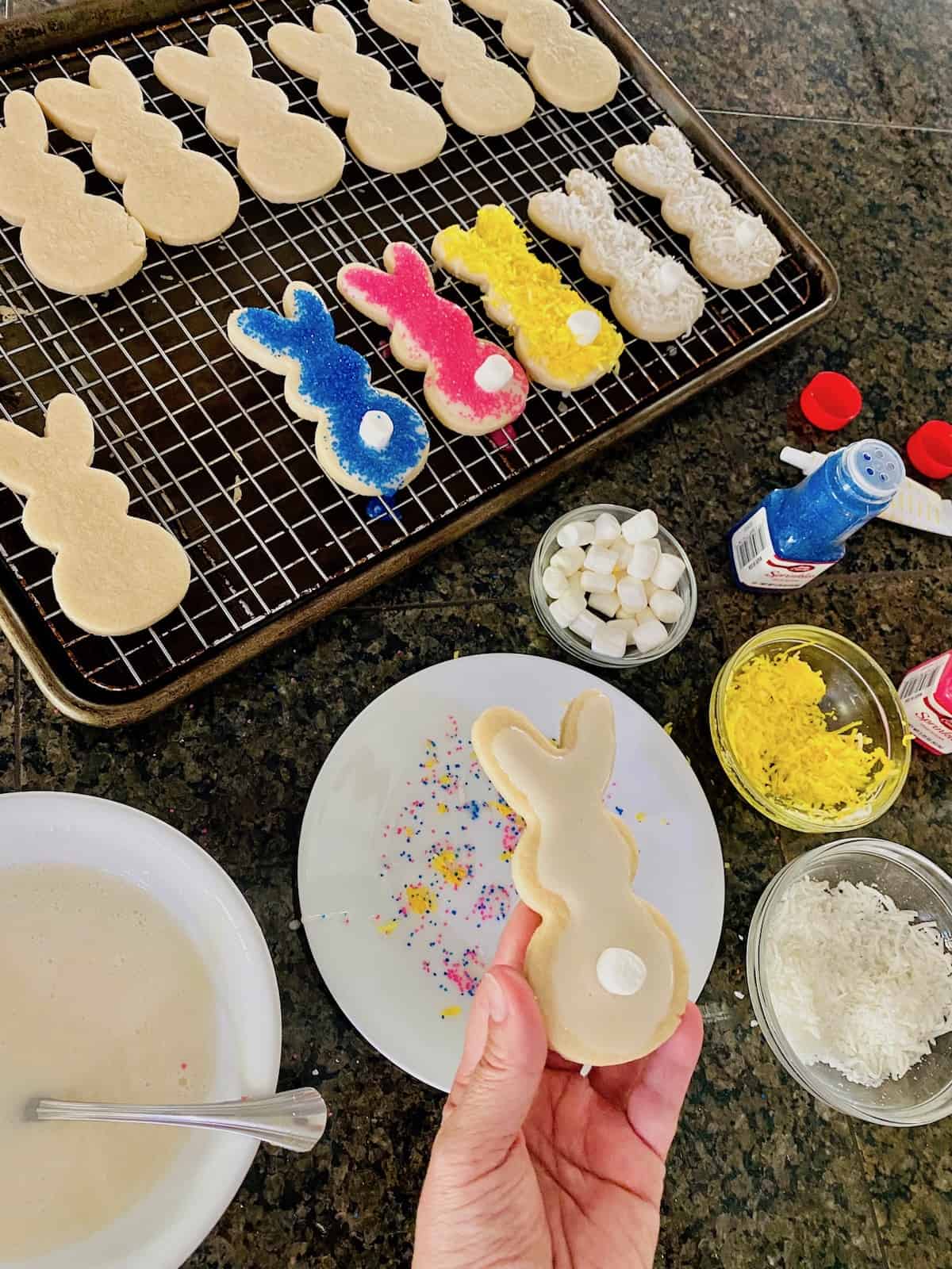 Baking sheet with some cookie plain and decorated plus hand holding a cooking with icing and a marshmallow as a bunny tail.