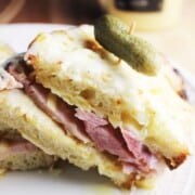 A sliced ham and cheese sandwich on a plate with a toothpick and pickle.
