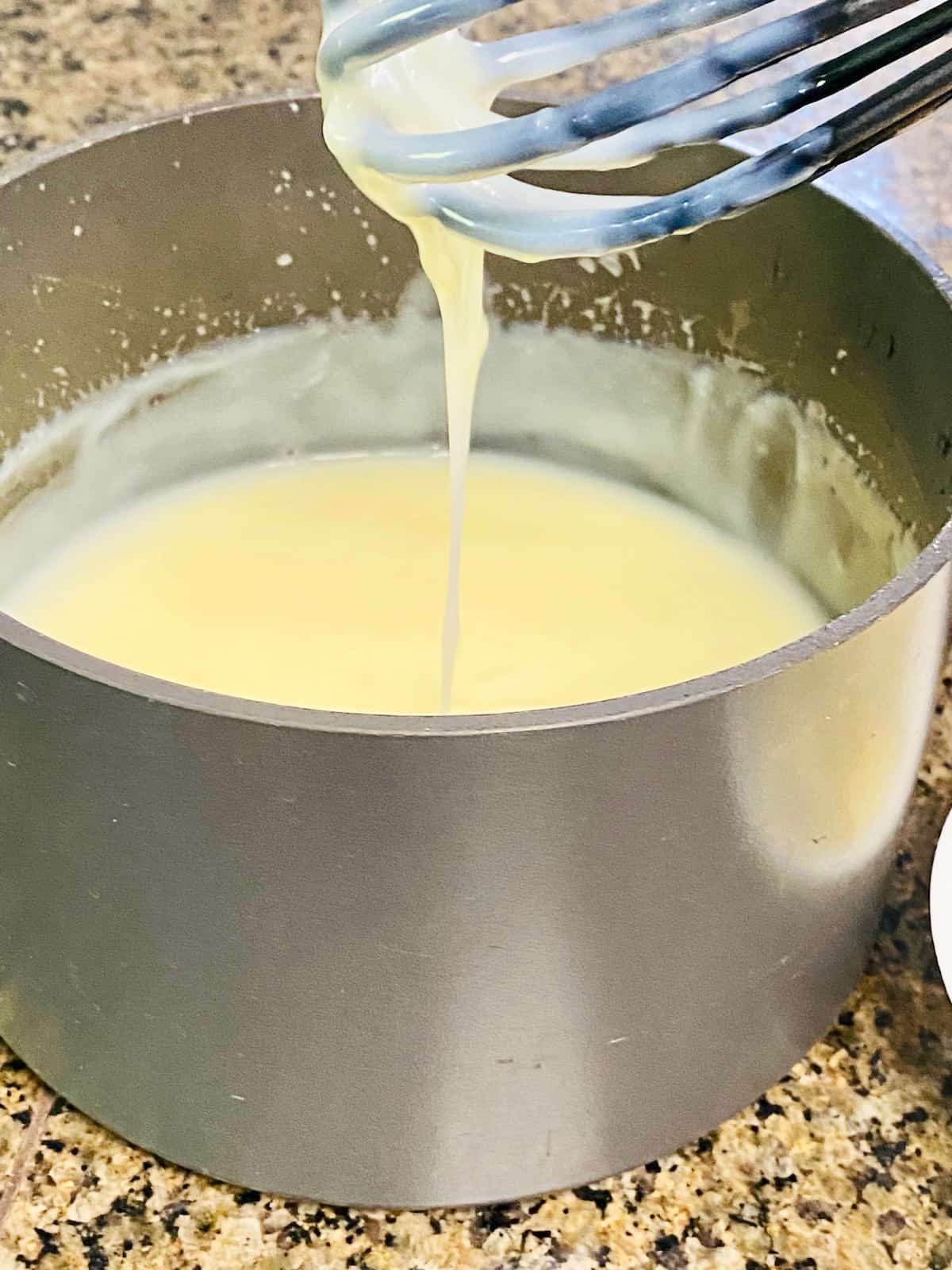 Whisk dipped into pot of cooked cream sugar custard showing thickness and a wide drip from whisk to pot contents.