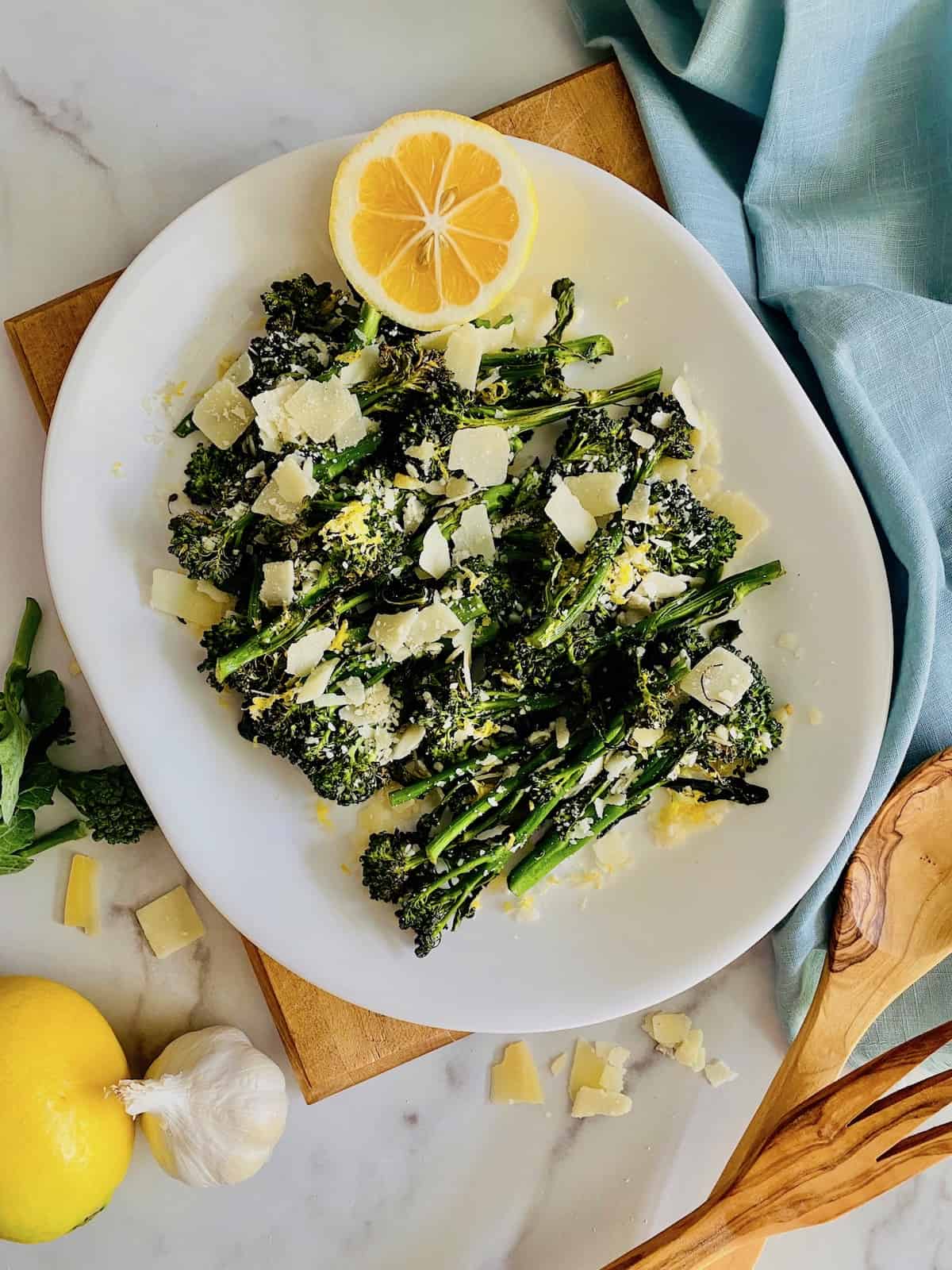Overhead of white platter filled with broccoli topped with parmesan and lemon juice and serving utensils and blue napkin to the side.