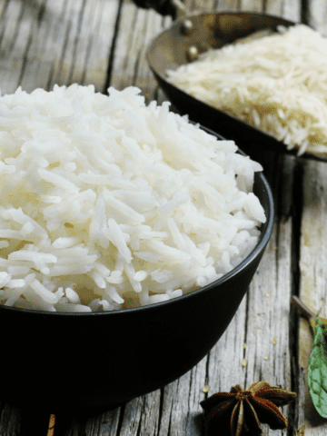 A bowl of cooked rice and a scoop with uncooked rice grains on a table.