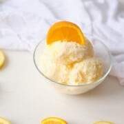 A glass bowl with 3 scoops of lemon orange ice cream topped with an orange wedge.