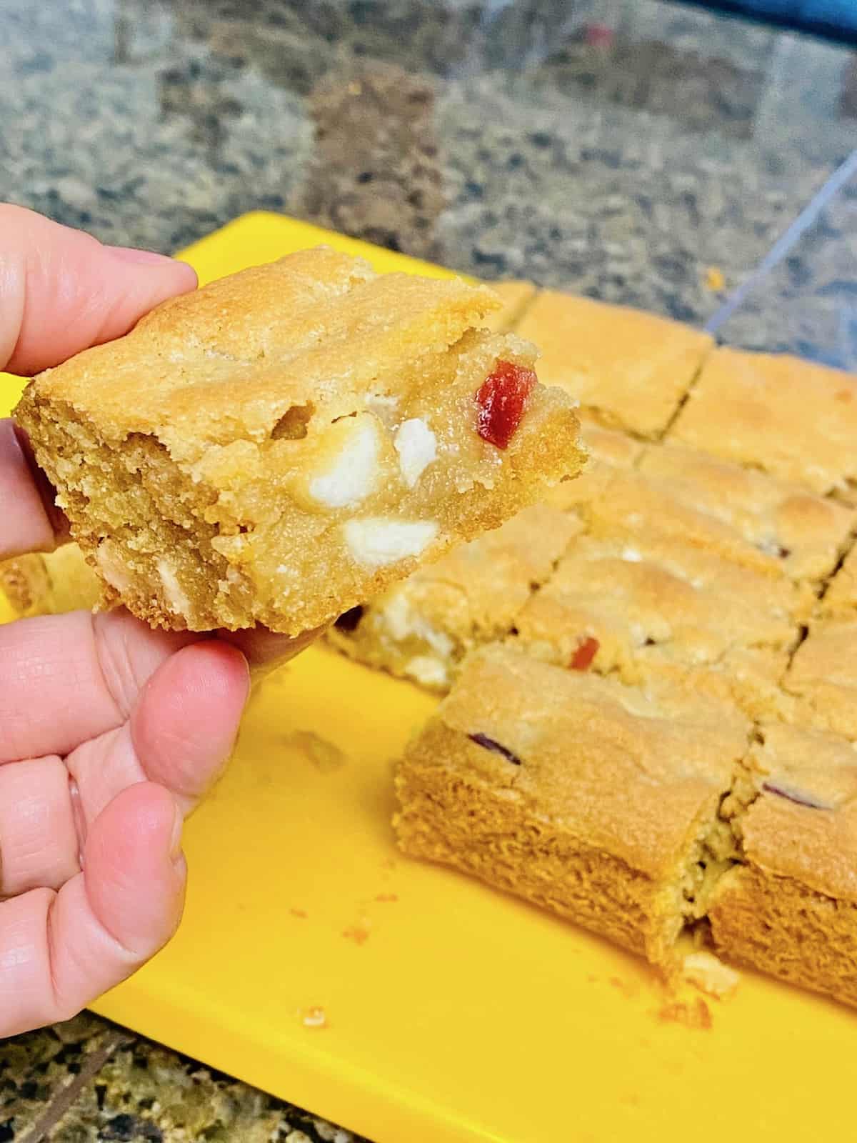 Strawberries & Cream Cookie Bars Cookies cut into squares on a cutting board and hand holding one close up to show the white chocolate chips and strawberries pieces inside.