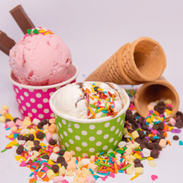 Two paper cups with scoops of ice cream with sprinkles on top and bunch of toppings all around on the table.
