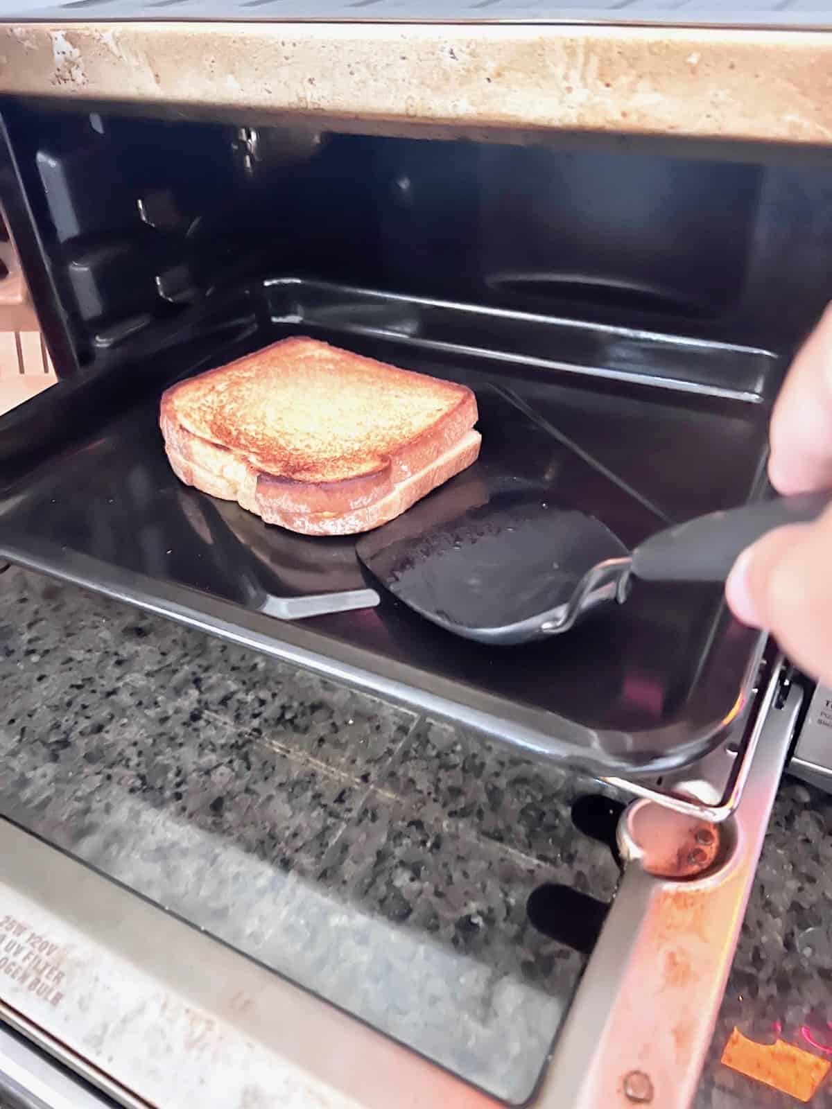 Toaster oven door open and using a spatula to flip the grilled cheese over.