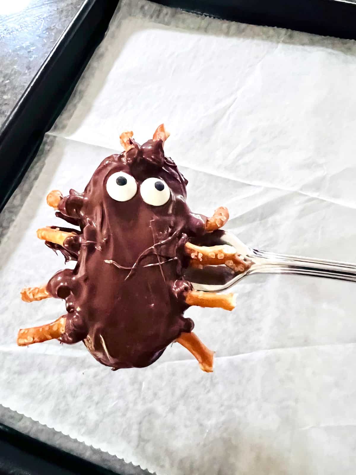 Fork holding a cookie dipped in chocolate with all the pretzel legs and antennae attached plus candy eyes.
