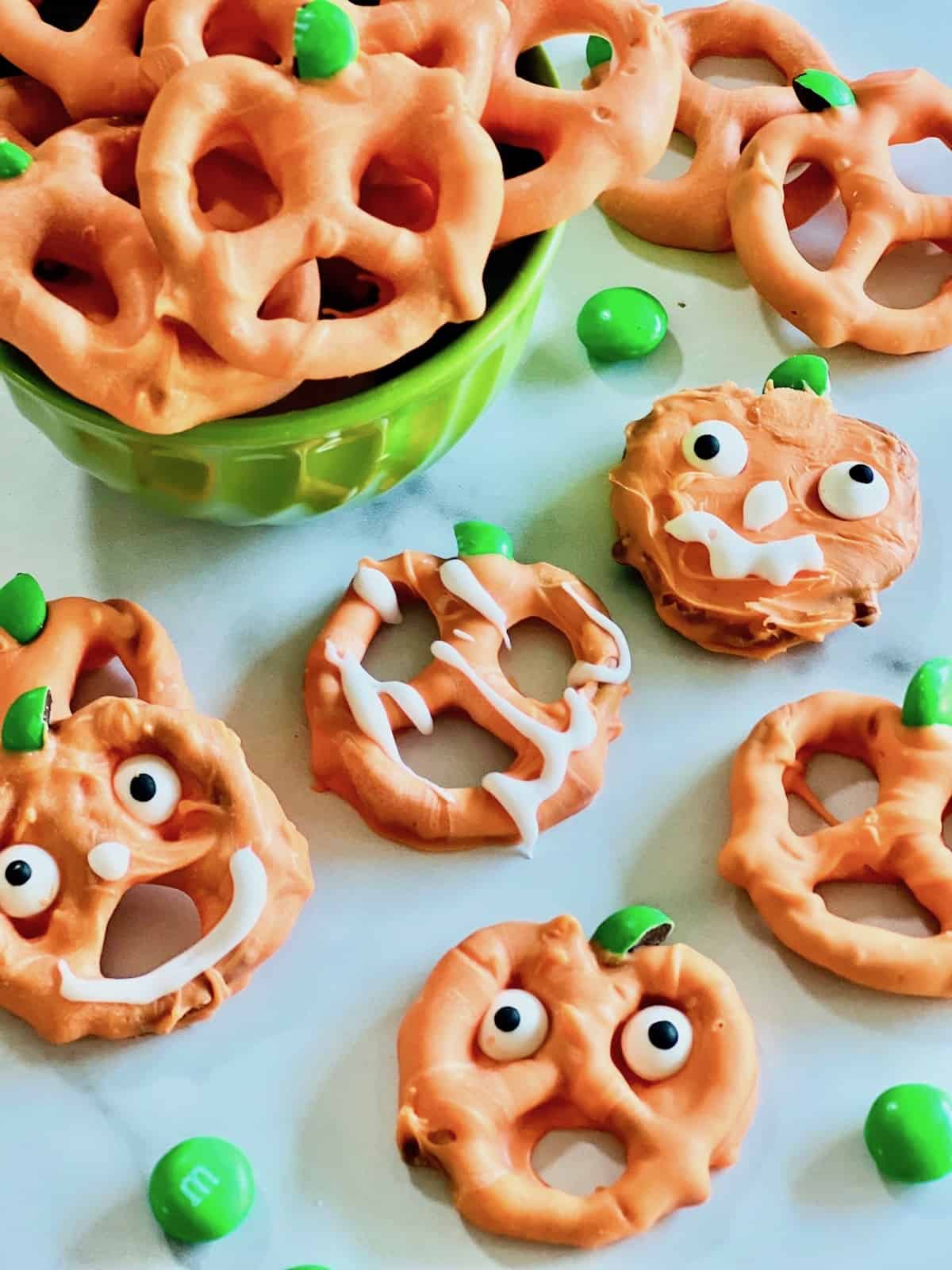 Pumpkin Pretzels Stacked in a green bowl with Jack O Lantern pretzels and M&Ms on the table.