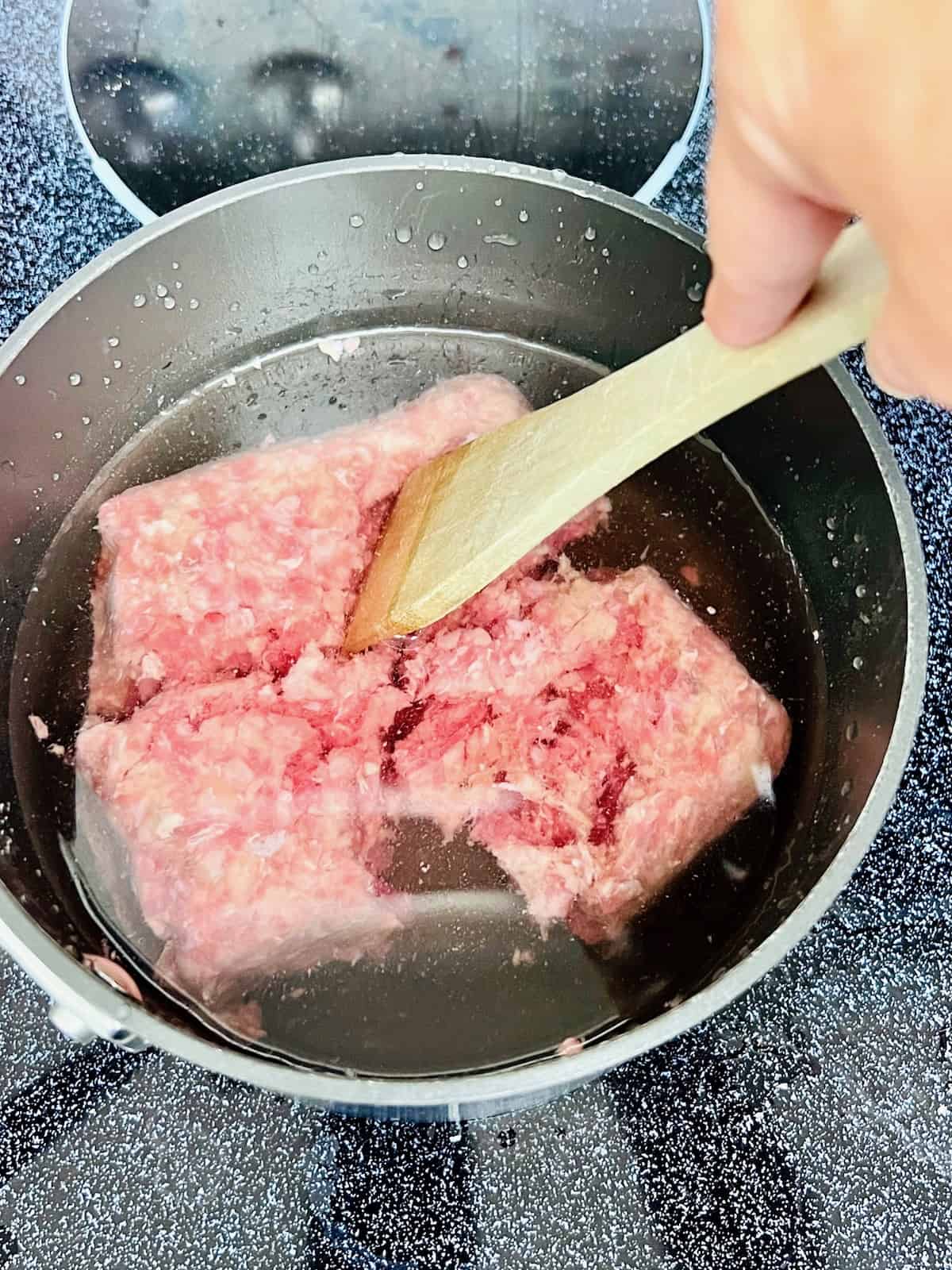 Ground beef in a pot covered with water and a wooden spatula breaking it apart.