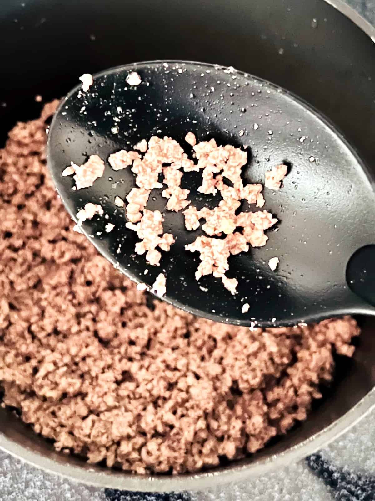 Ground beef in the pot cooked and a spoon showing finely ground pieces.