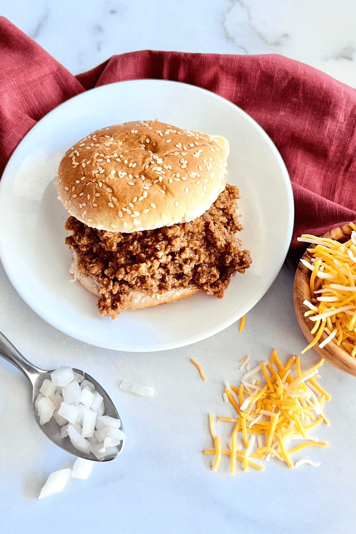 3-Ingredient Sloppy Joes (Gluten-Free) On a bun with shredded cheese and chopped onions to the side for topping.