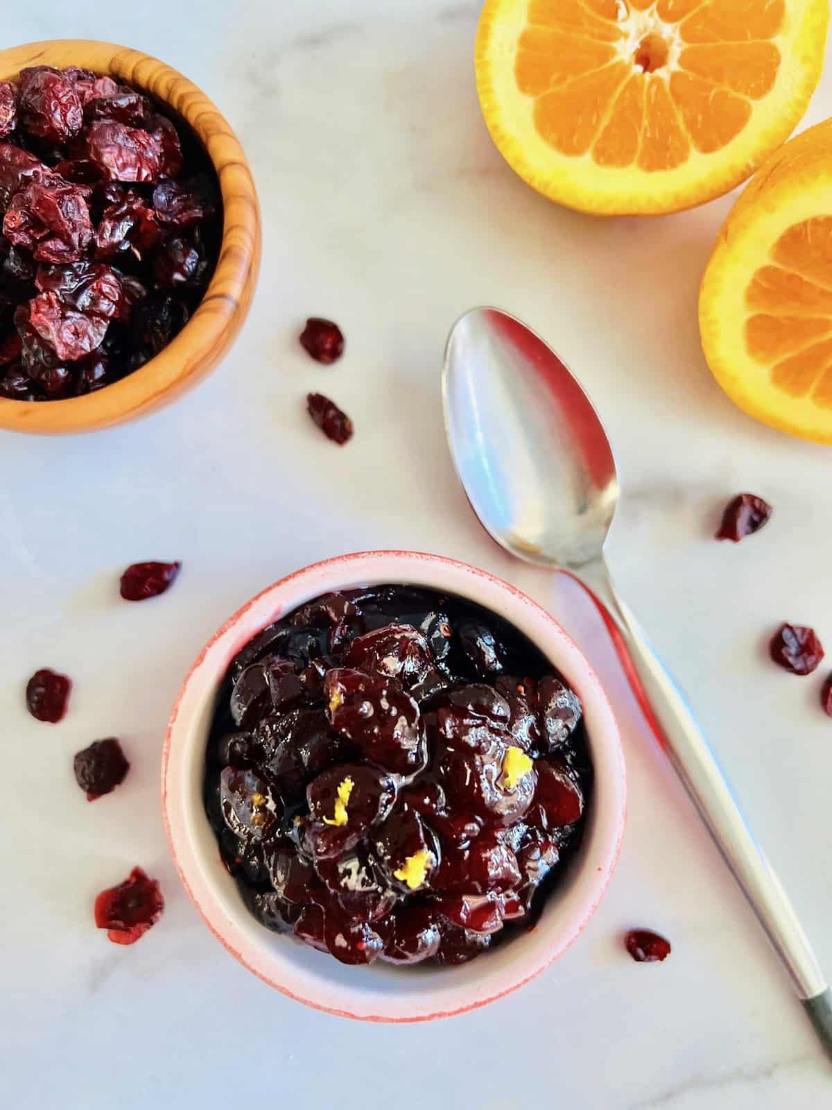 Cranberry sauce in a small bowl ready to eat with a spoon surrounded by a bowl of dried cranberries and a fresh orange.