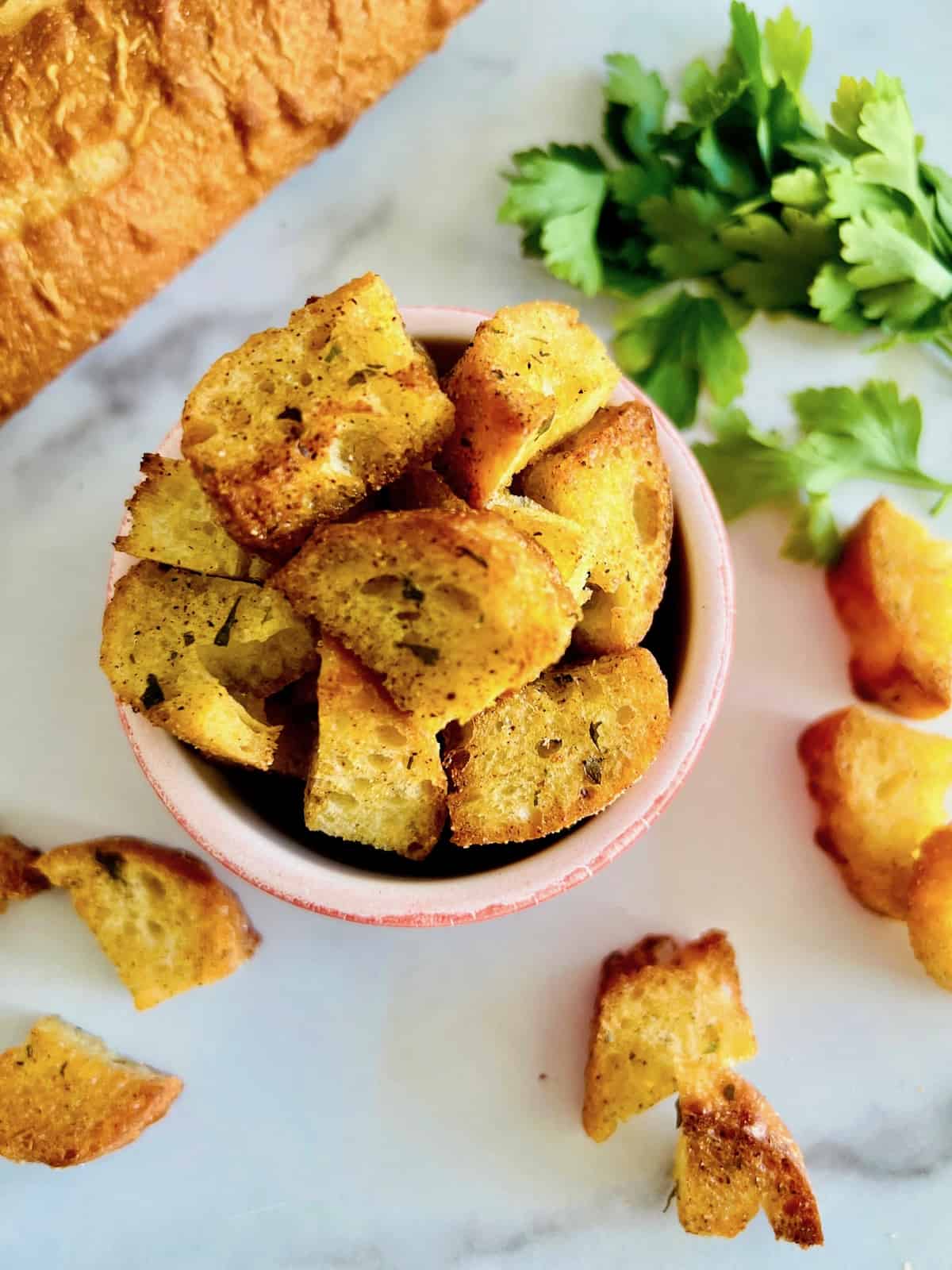  Overhead image of air fryer croutons freshly toated in a bowl with a baguette croutons, and parsley on the table.
