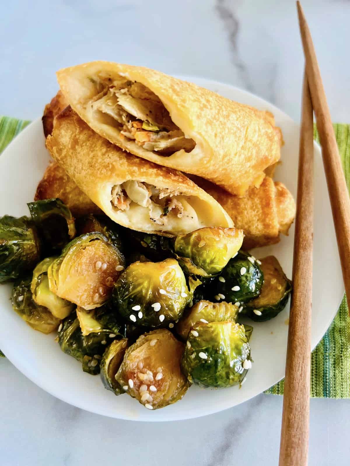 Asian-Inspired Teriyaki Brussels Sprouts Plated with air fried egg rolls on top & chopsticks.