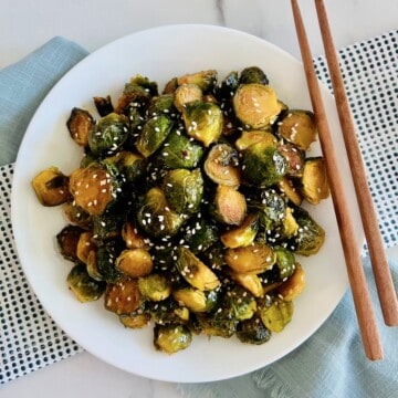 Asian-Inspired Teriyaki Brussels Sprouts topped with sesame seeds plated with a cloth napkin and chopsticks.