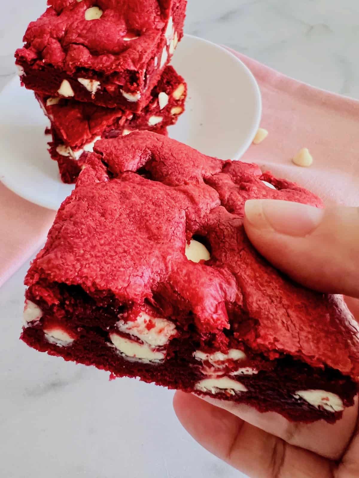 Red Velvet Brownies with Cake Mix Hand holding one and a plate of them in the background.