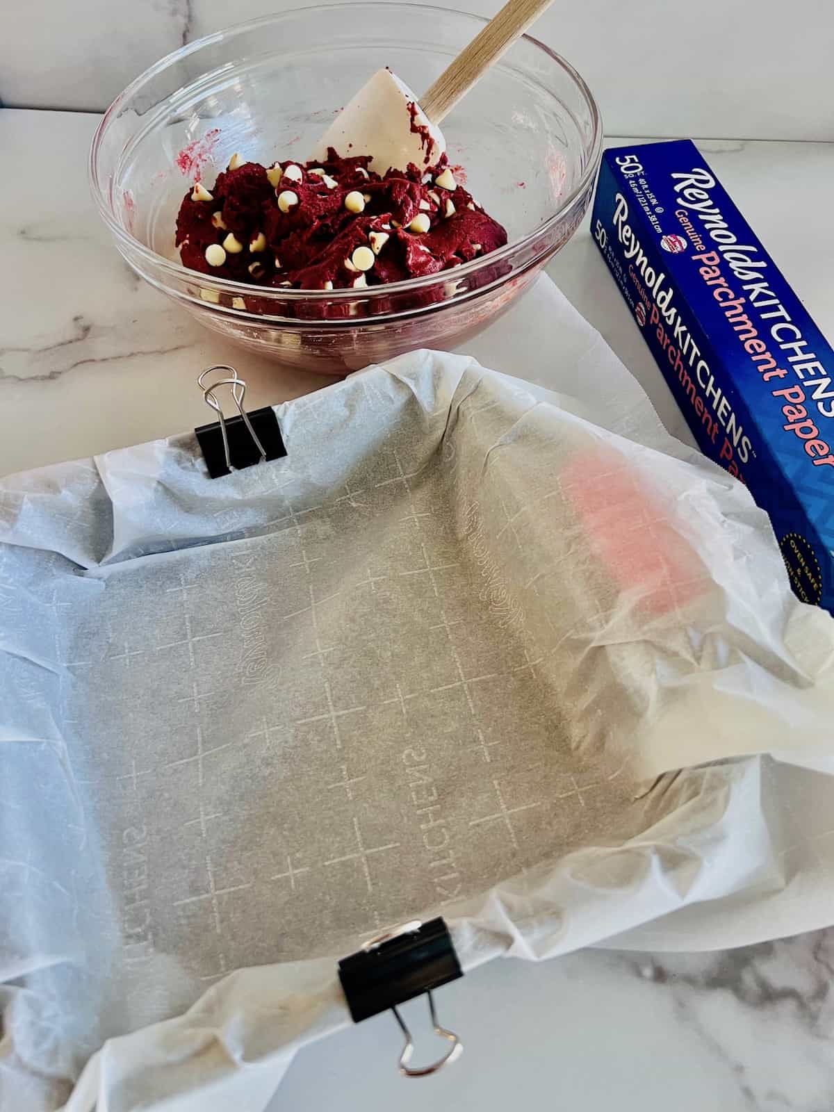 Red Velvet Brownies with Cake Mix parchment lined pan held with binder clips & red velvet brownie batter in a bowl.