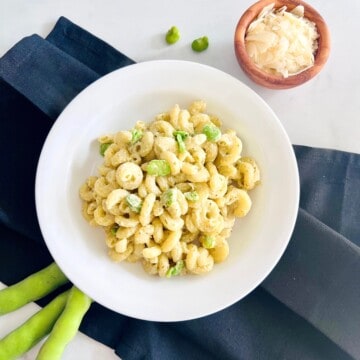 White pasta bowl filled with cavatappi pasta in a creamy pesto sauce topped with fava beans & parmesan cheese.