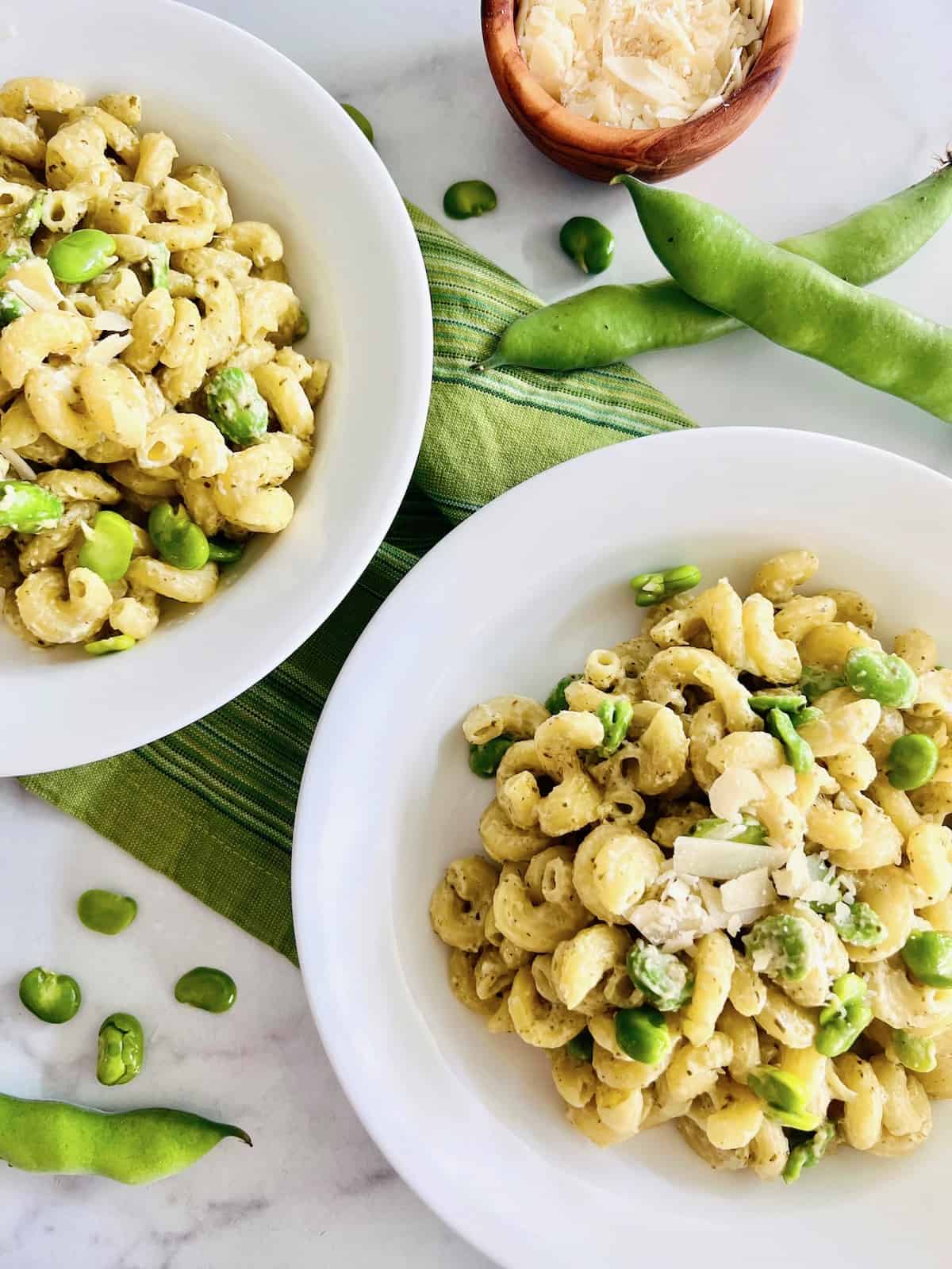 Creamy Pesto Pasta with Fava Beans Two pasta bowls filled with shaved parmesan on top and fava beans with green napkin.