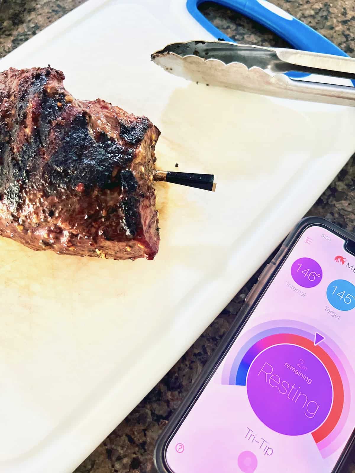 Grilled Tri-Tip on the cutting board with the Meater prob sticking out and the app open on a phone showing it is resting.