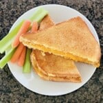 Overhead image of a grilled cheese sandwich fresh from the microwave cut in half on plated with carrots and celery.