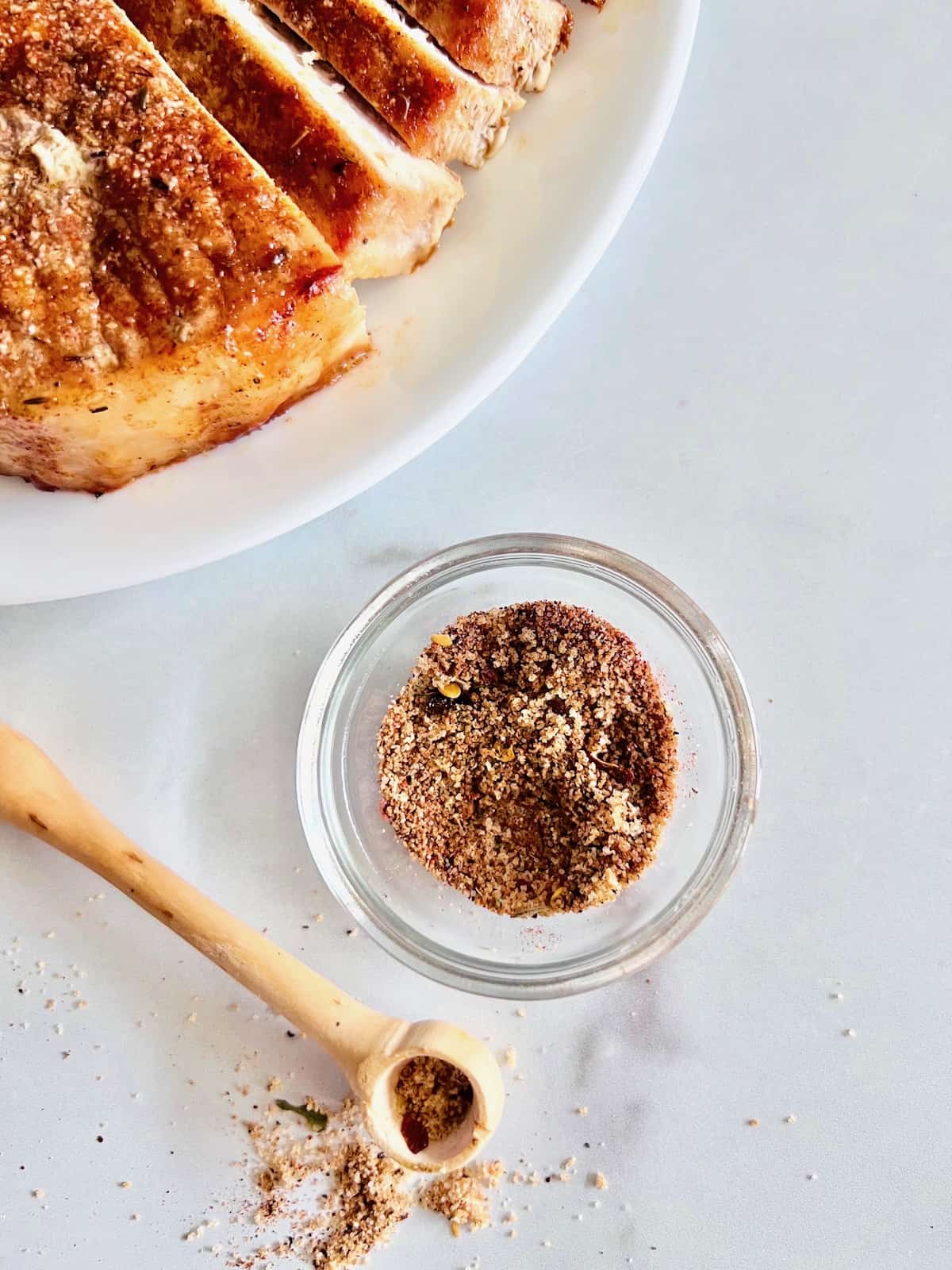 Dry Rub for Pork Chops Glass bowl of dry rub with wooden spice spoon spilling out next to plate with sliced pork boneless chop,