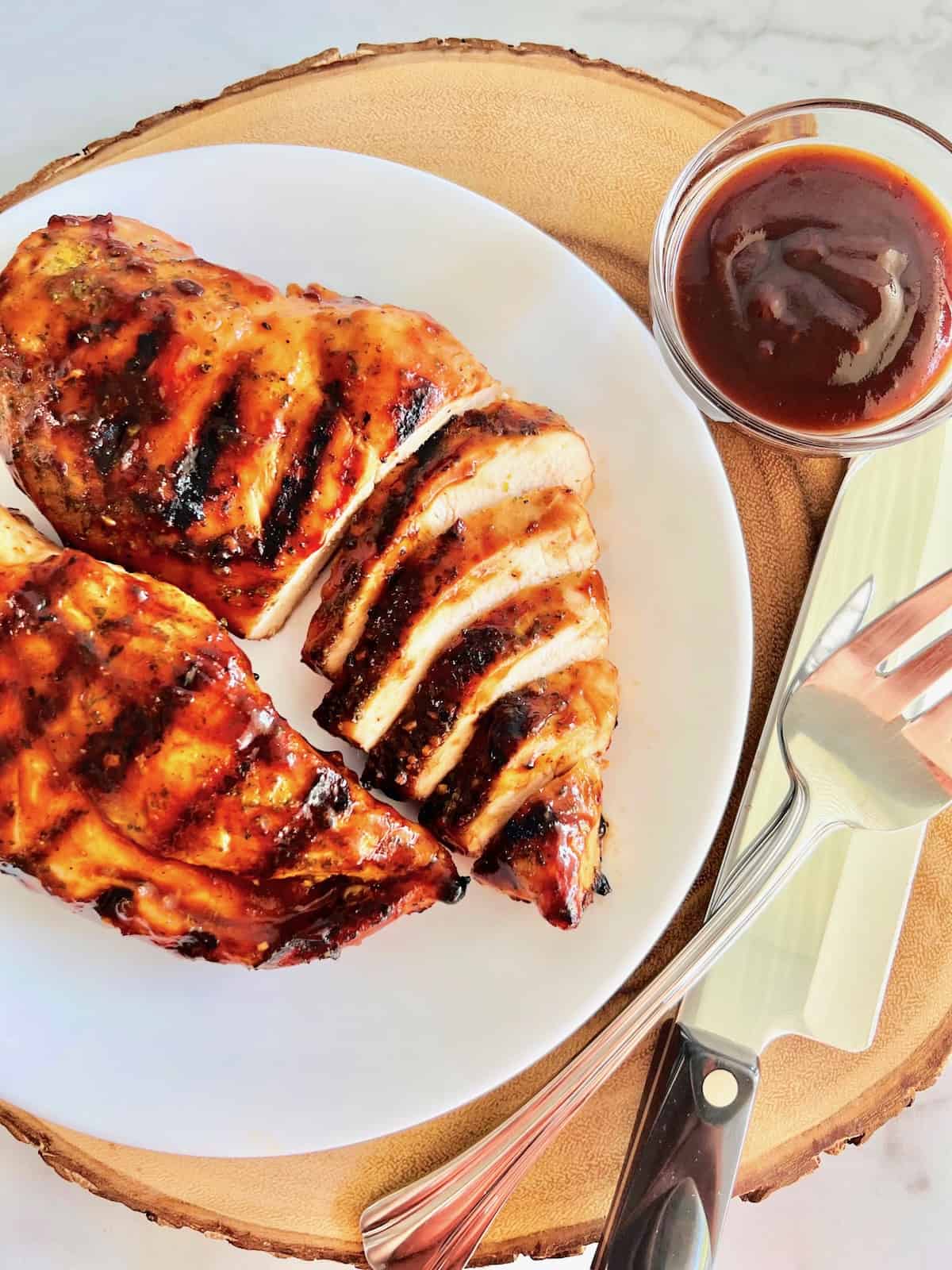 Grilled BBQ Chicken Breasts Served on a plate with some slices plus bowl of bbq sauce and utensils on the side.