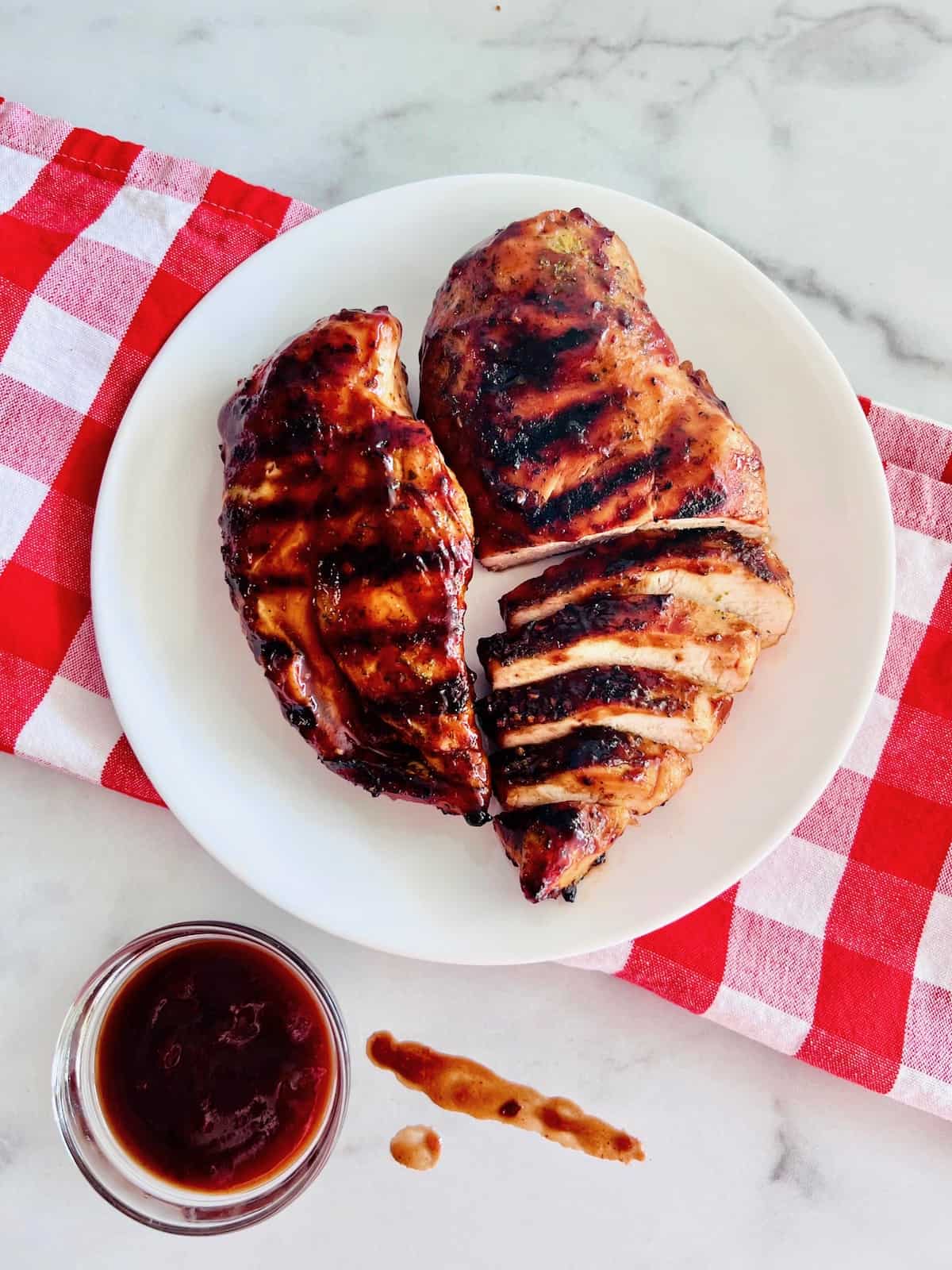 Grilled BBQ Chicken Breasts Sliced & whole on a plate with bbq sauce in a dipping bowl and some smeared on the counter.
