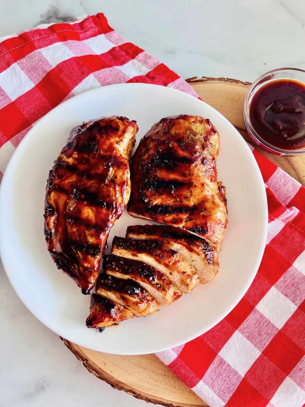 Grilled BBQ Chicken Breasts ready to serve plated on a red checker napkin with extra bbq sauce in a bowl.