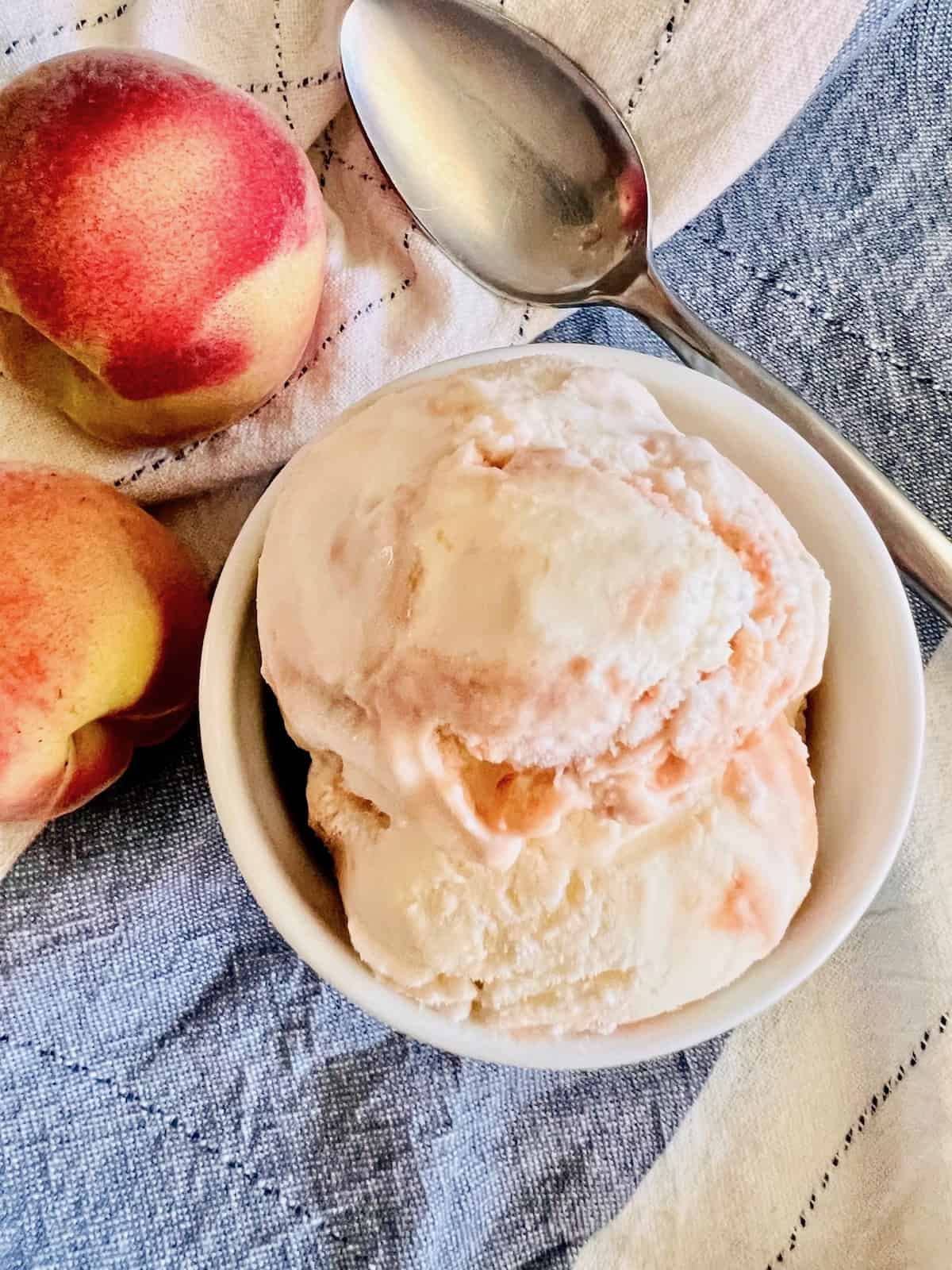 Peaches & Cream Ice Cream A scoop in a white ramekin next to a spoon and peaches on top of a cloth napkin.