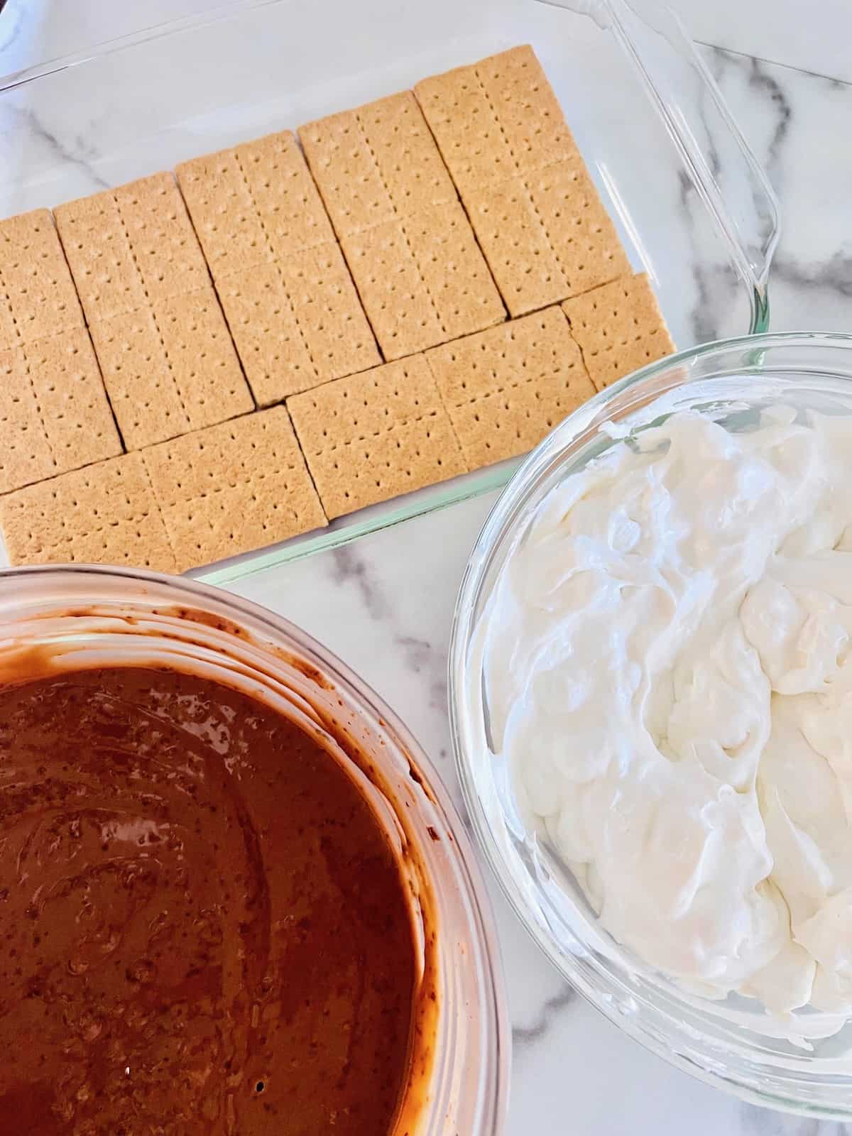 S’mores Icebox Cake Pan lined with graham crackers and bowls filled with the chocolate pudding and marshmallow cream.