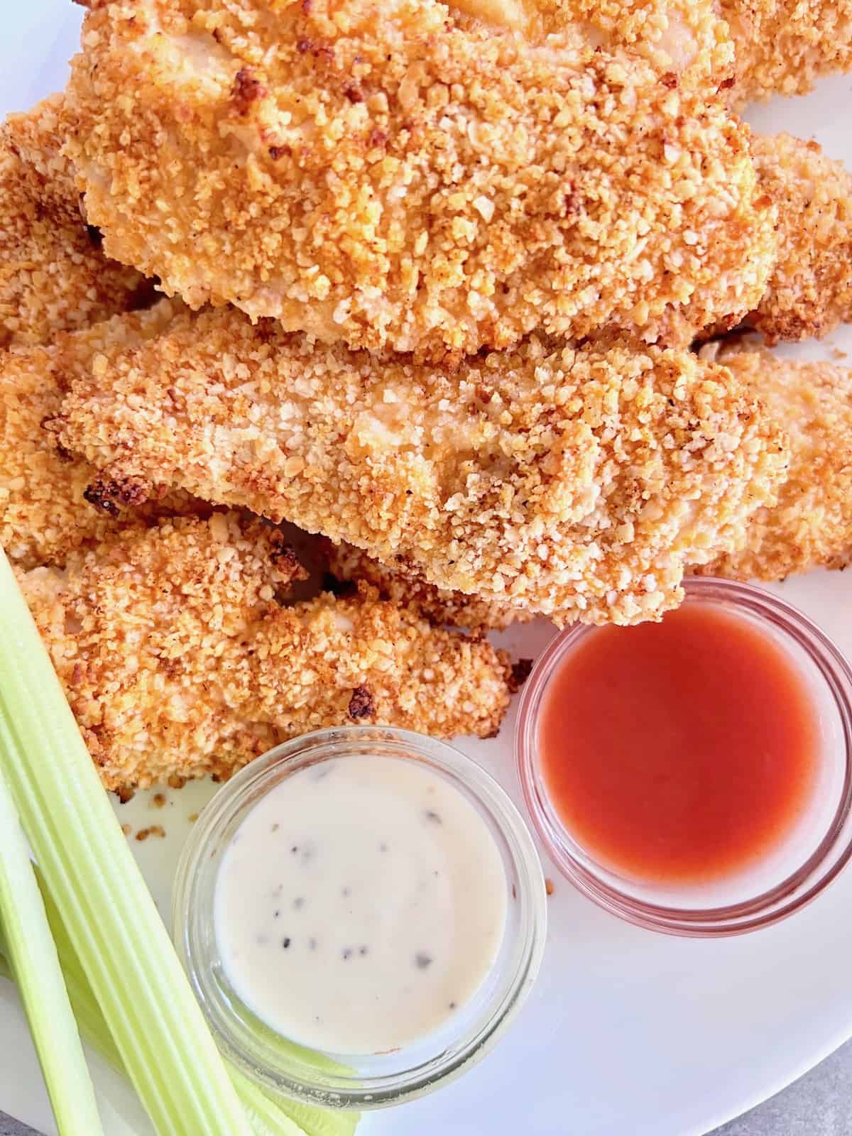 Air Fryer Buffalo Chicken Tenders Closeup stacked on a white plate with dips on the bottom & celery to the left.