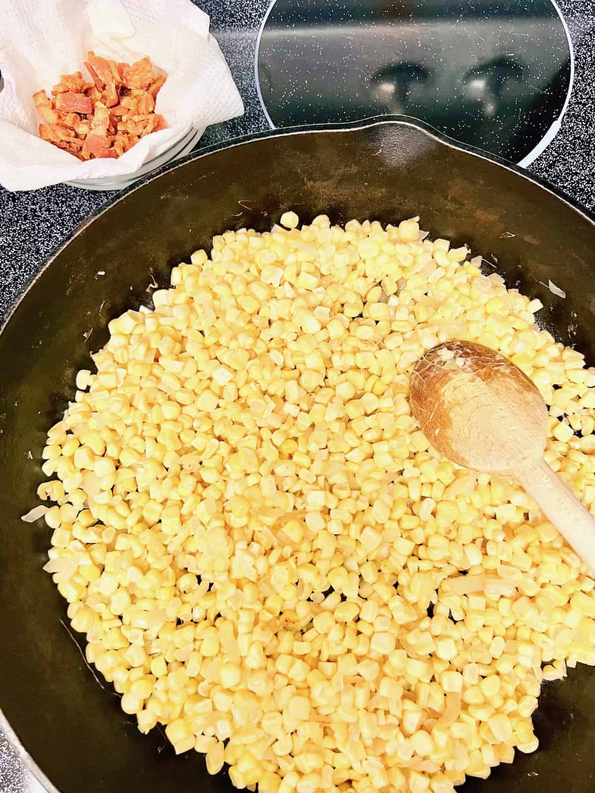 Stirring corn in the skillet next to bacon cooked in a paper towel lined bowl.