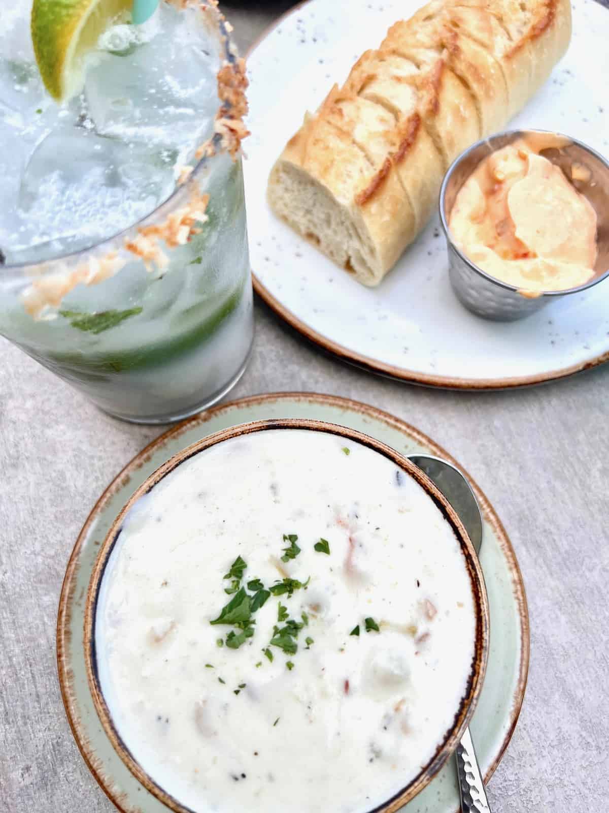 Cup of clam chowder next to fresh sliced bread, butter, & a coconut mojito cocktail.