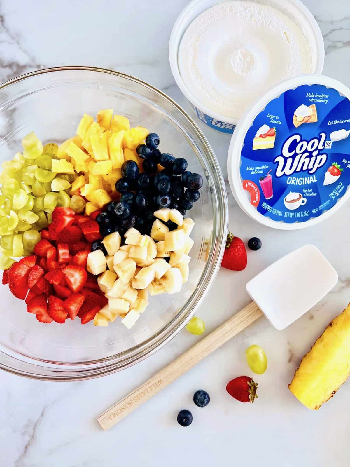 Fresh fruit blueberries and cut grapes strawberries pineapple and banana in a mixing bowl next to container of cool whip.