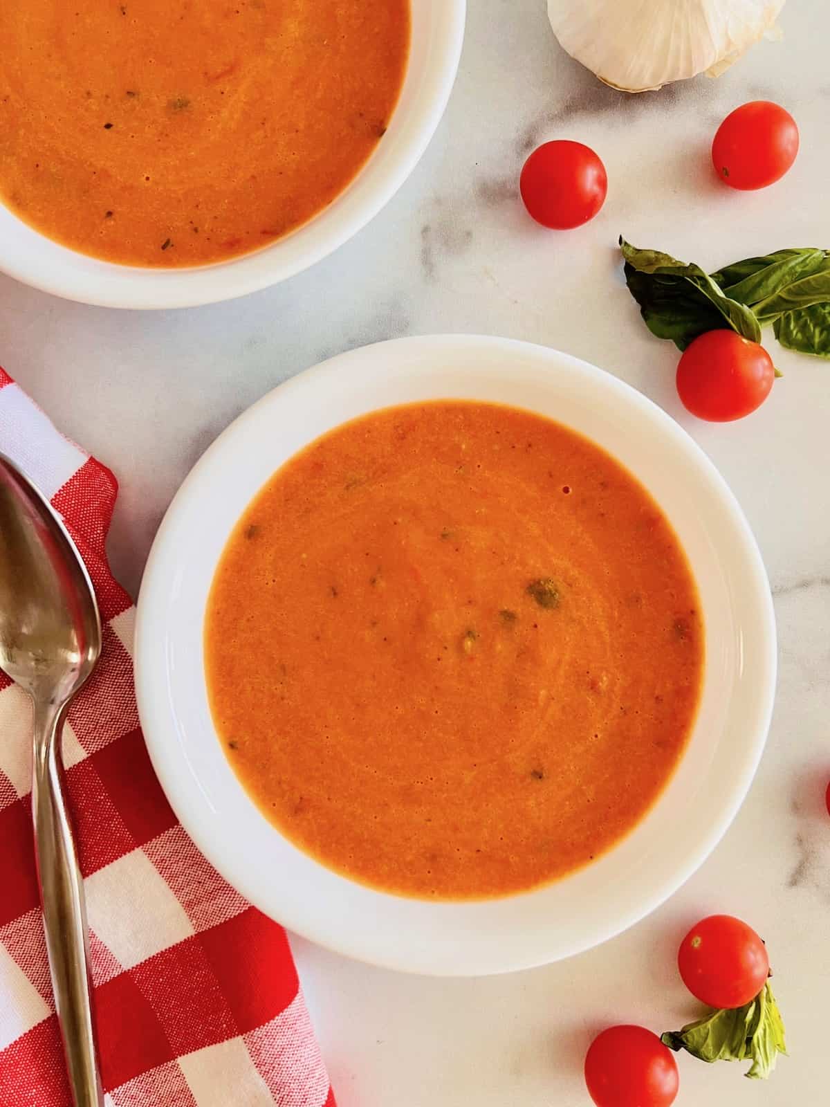 Overhead two bowls of tomato soup next to cherry tomatoes spoon napkin and basil leaves.
