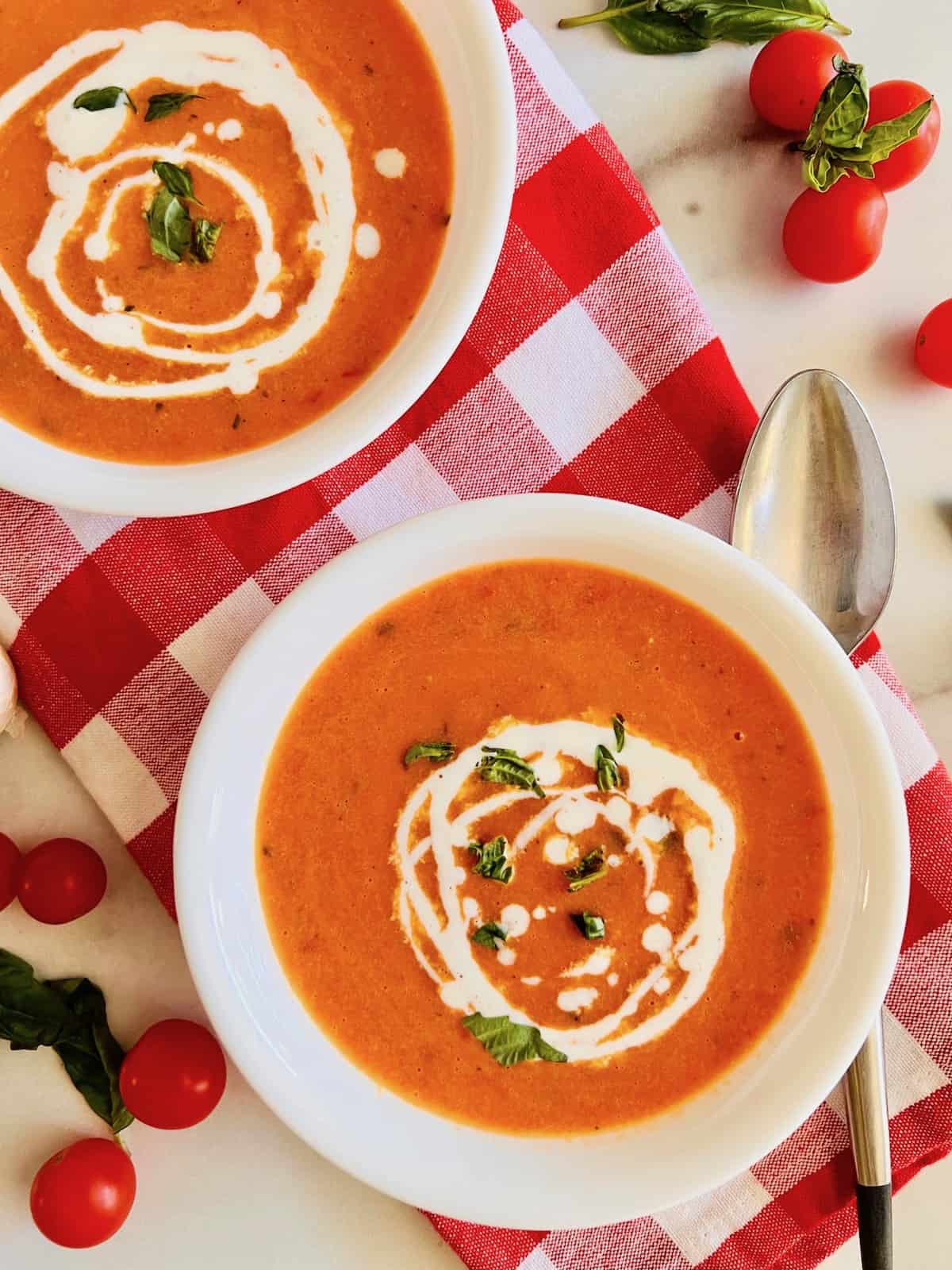Roasted Cherry Tomato Soup Overhead two bowls topped with fresh basil leaves and swirls of cream on red checkered napkin.